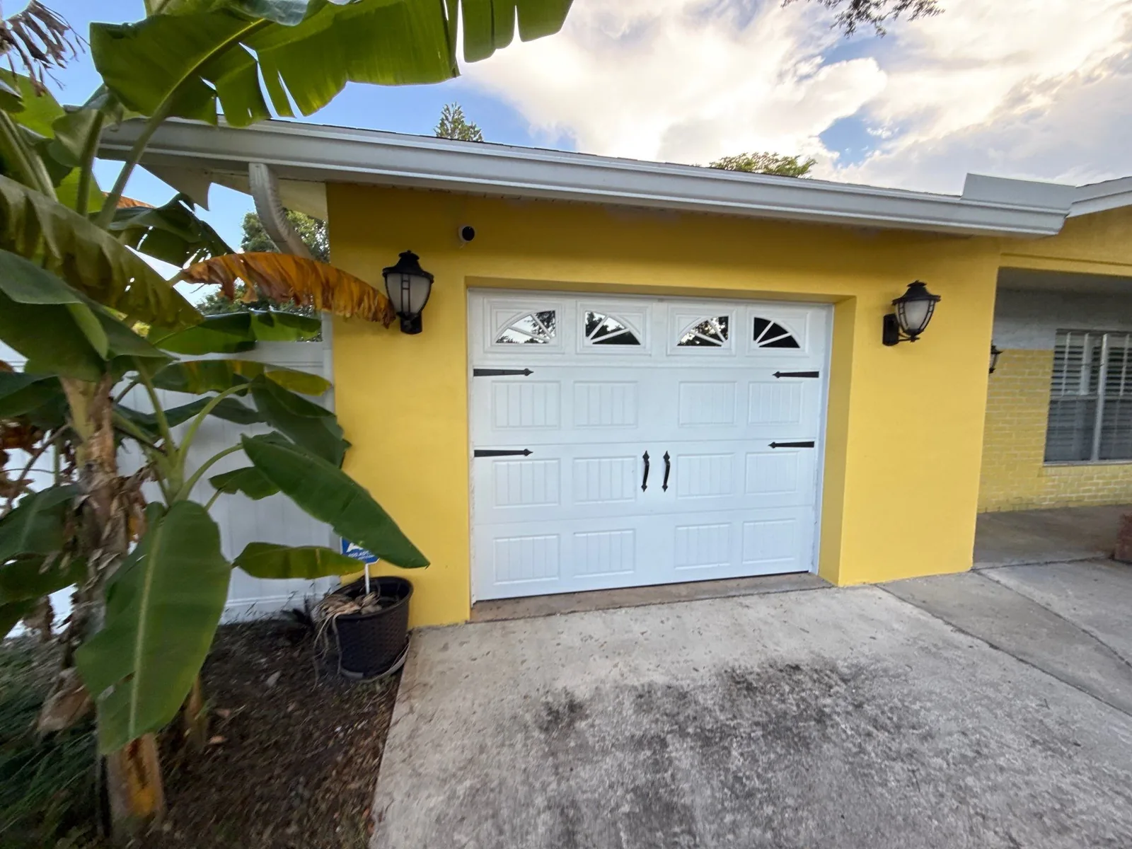 Exterior Painting in River Park, Florida. Yellow stucco home exterior with a white carriage-style garage door, white fascia and black coach lights, with a banana plant in the foreground.