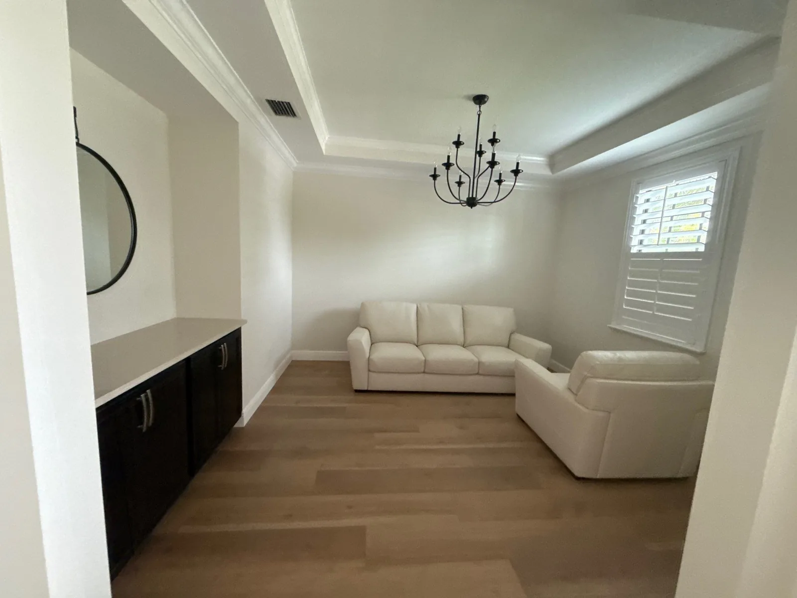 Interior Painting in Royal Palm Beach, Florida. Sitting room with white walls and a white tray ceiling with crown moulding, black chandelier, plantation shutters and two cream sofas.