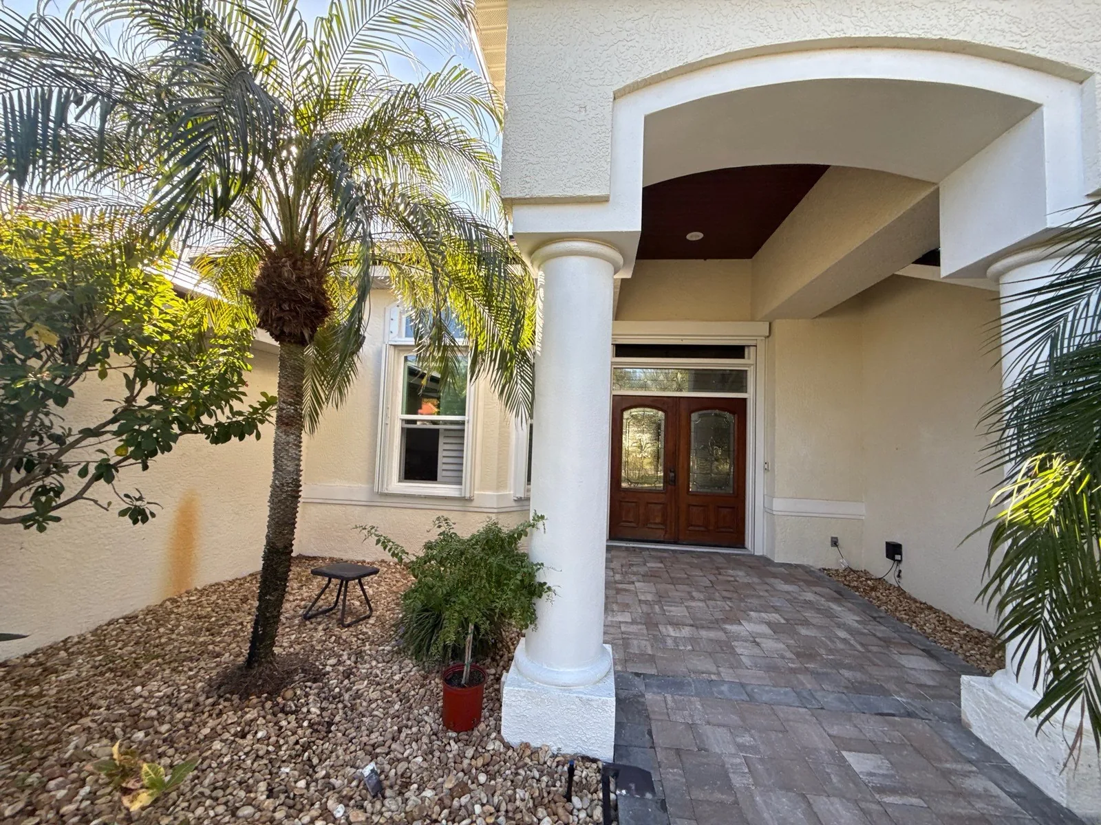 Exterior Painting in Satellite Beach, Florida. Covered front entrance with white column, cream stucco walls and dark wood double doors, paver walkway and palm.