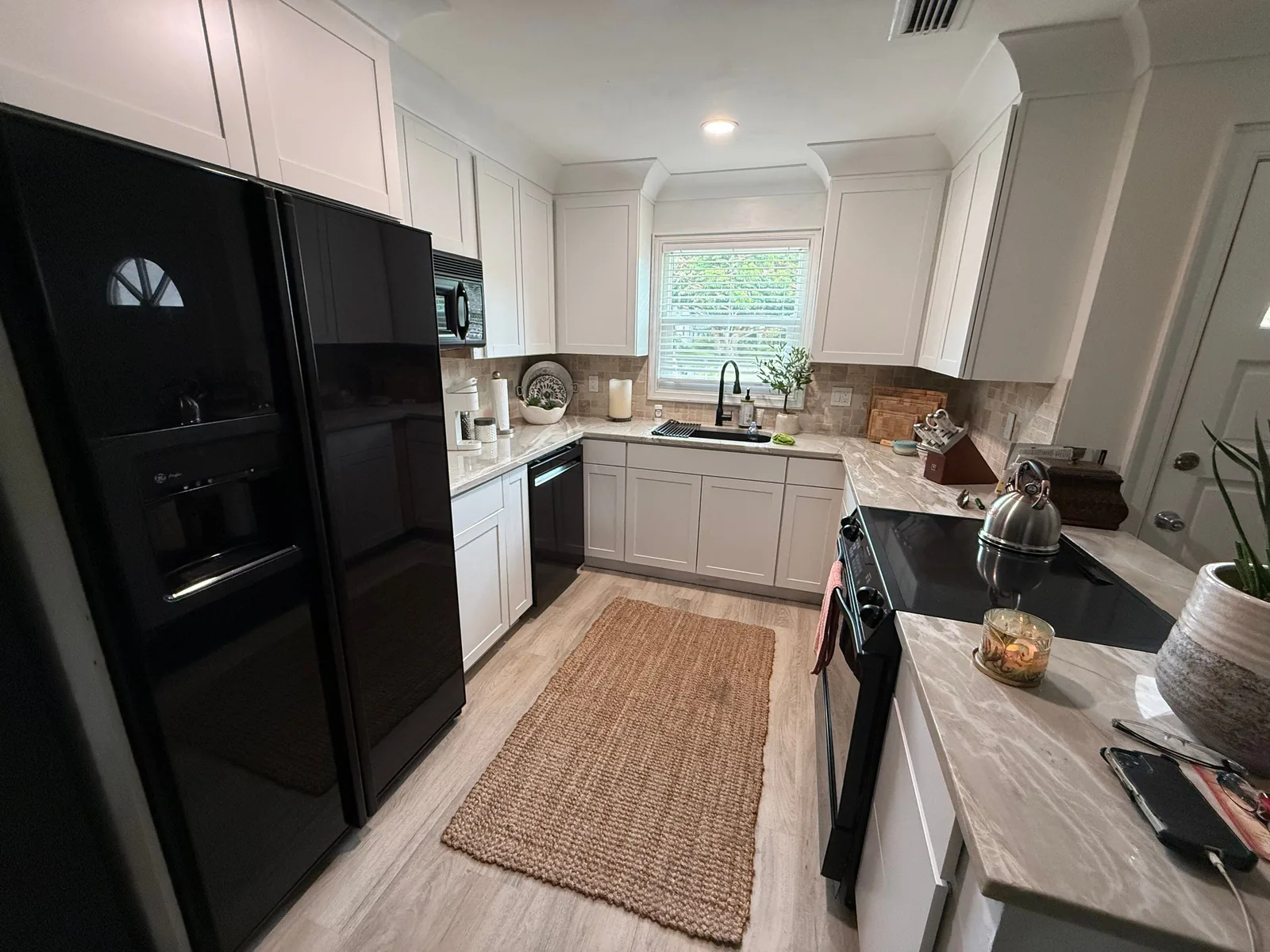 Cabinet Refinishing in Sebastian, Florida. Kitchen with white painted upper cabinets and light grey lower cabinets, black refrigerator and dishwasher, light stone countertops and a tile backsplash.