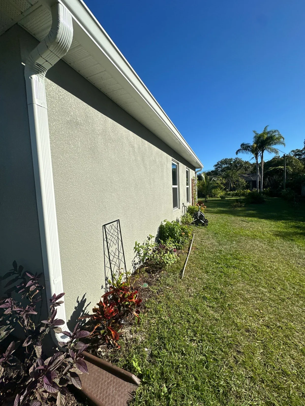 Exterior Painting in Sebastian, Florida. Side elevation of a single-storey stucco house in pale grey with white gutter and fascia, with planting beds and lawn under a clear blue sky.