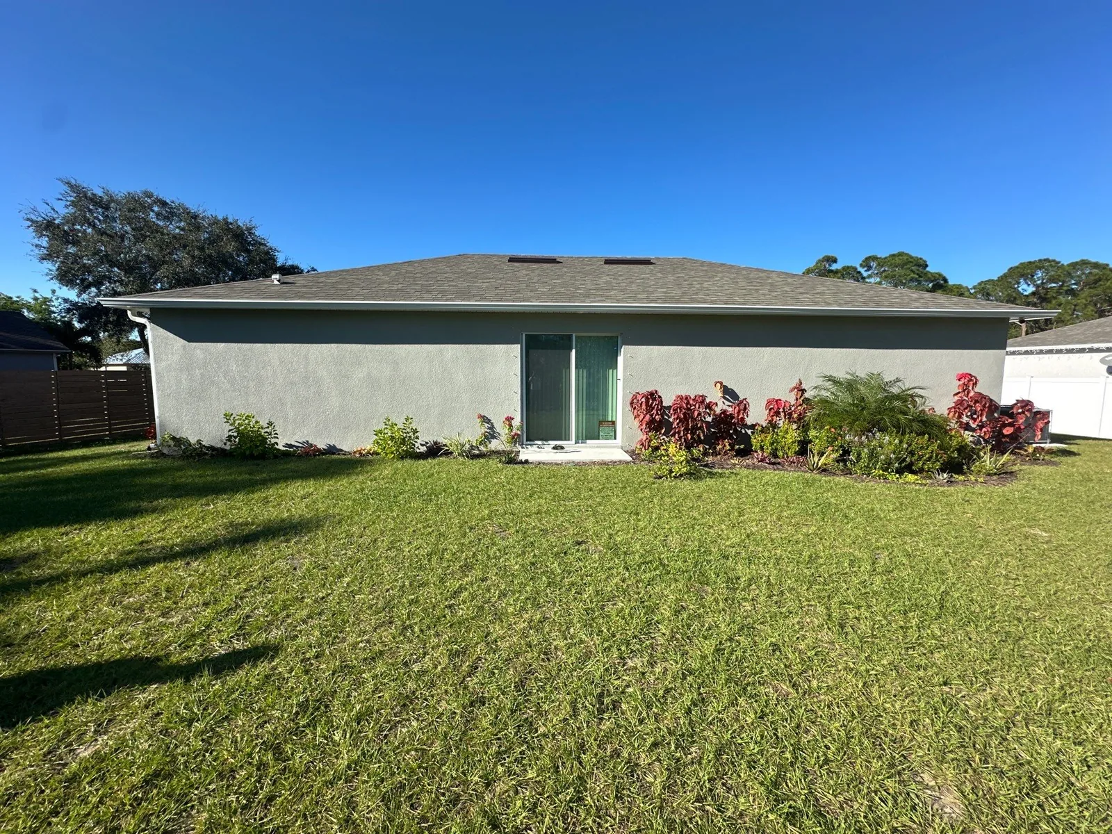 Exterior Painting in Sebastian, Florida. Rear elevation of a single-storey stucco house in grey with a sliding glass door, planting beds and mown lawn under a clear sky.