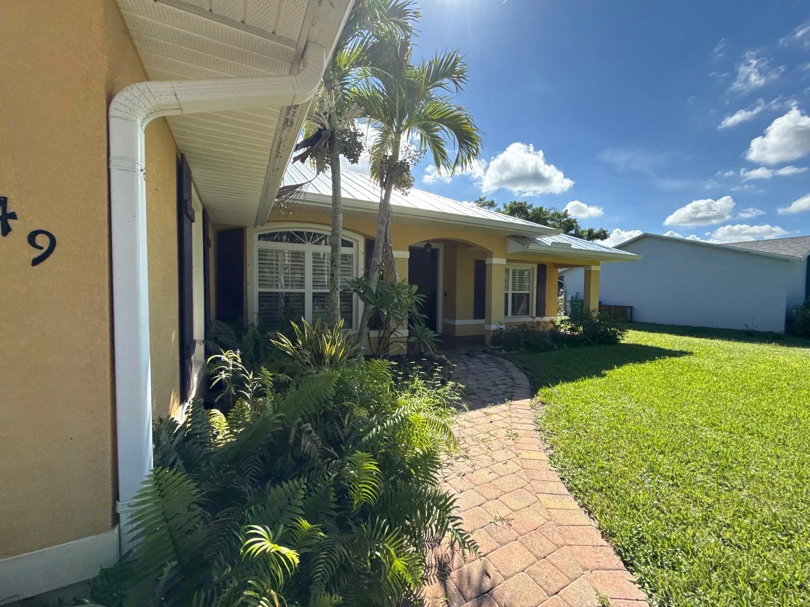 Exterior Painting in Sebastian, Florida. Single-storey home with yellow stucco walls, white fascia and trim and dark shutters, brick paver walkway leading past palms and lawn.