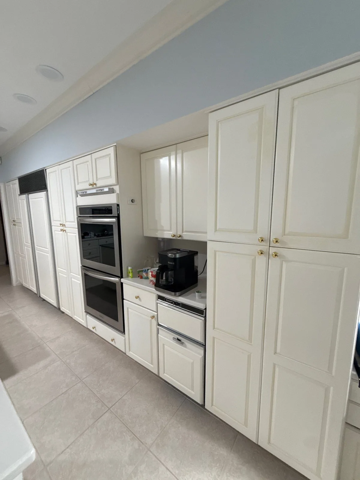Cabinet Refinishing in South Beach, Florida. Run of off-white raised-panel kitchen cabinets with wall oven and pantry doors, tile floor.