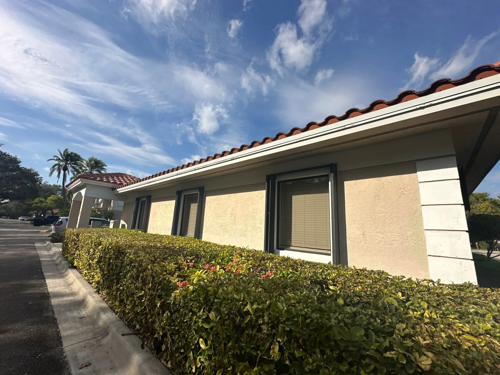 Exterior Painting in Stuart, Florida. Single-storey stucco building exterior, tan walls with dark window frames and a barrel-tile roof, behind a trimmed hedge.