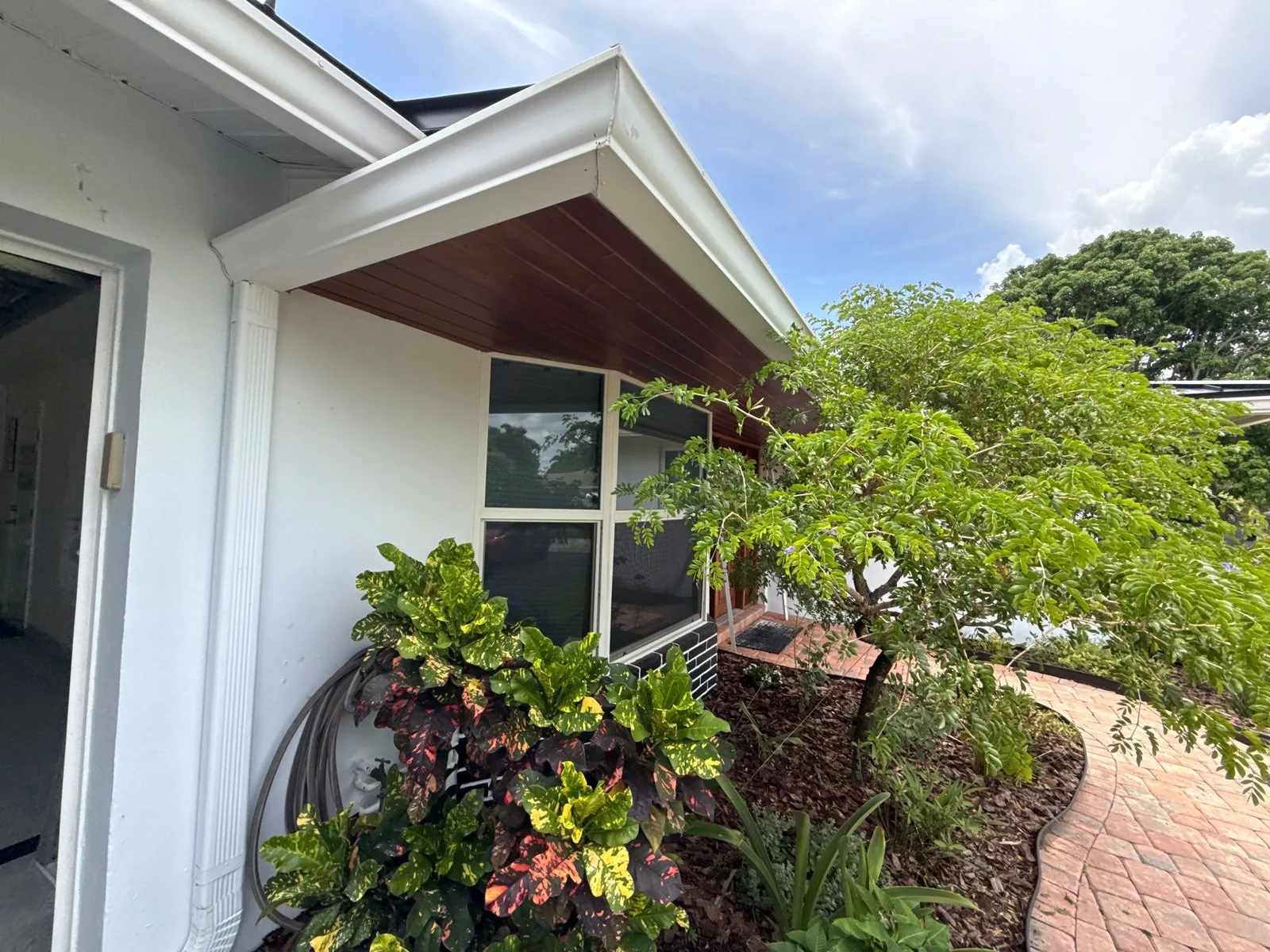 Exterior Painting in Tequesta, Florida. Side elevation of a white-painted house with a stained wood eave overhang, picture window and planted bed.