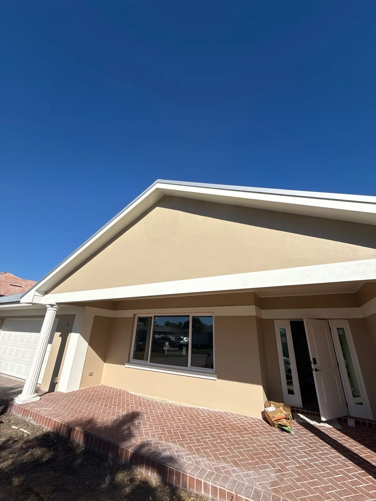 Exterior Painting in Vero Beach, Florida. Front entry of a single-storey home with tan stucco gable, white fascia and trim, dark front door and brick paver entry.