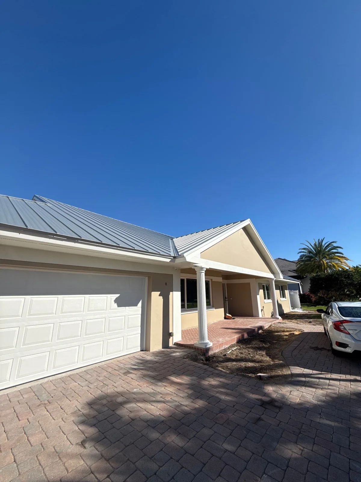 Exterior Painting in Vero Beach, Florida. Front of a single-storey home with tan stucco, white trim, a standing-seam metal roof, white garage door and paver driveway.