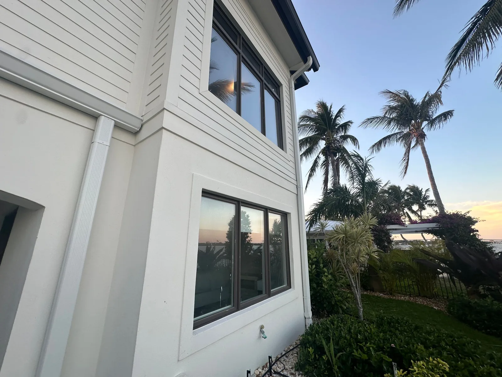 Exterior Painting in Vero Beach, Florida. Corner of a white lap-siding two-storey home with dark-framed windows, palm trees and water beyond at dusk.