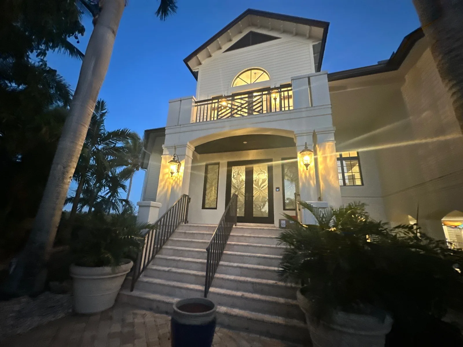 Exterior Painting in Vero Beach, Florida. Two-storey coastal-style home exterior painted light with white trim, balcony, lit wall sconces and a stone entry staircase.