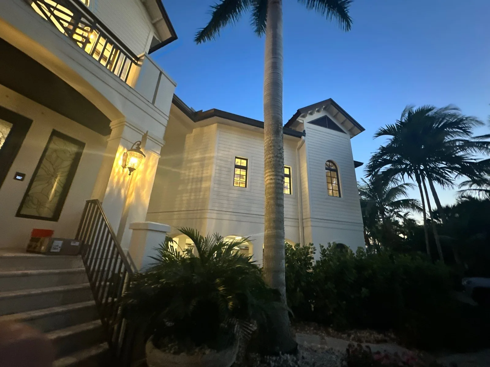 Exterior Painting in Vero Beach, Florida. Two-storey white home at dusk with exterior lights on, an entry staircase with a dark metal railing and palm trees.