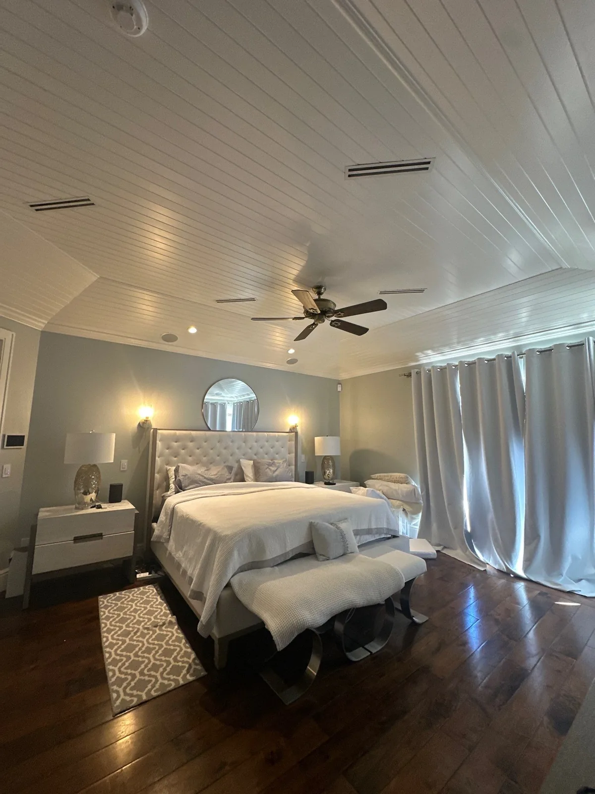Interior Painting in Vero Beach, Florida. Furnished bedroom showing white plank vaulted ceiling with ceiling fan, pale blue-grey walls and floor-length curtains.