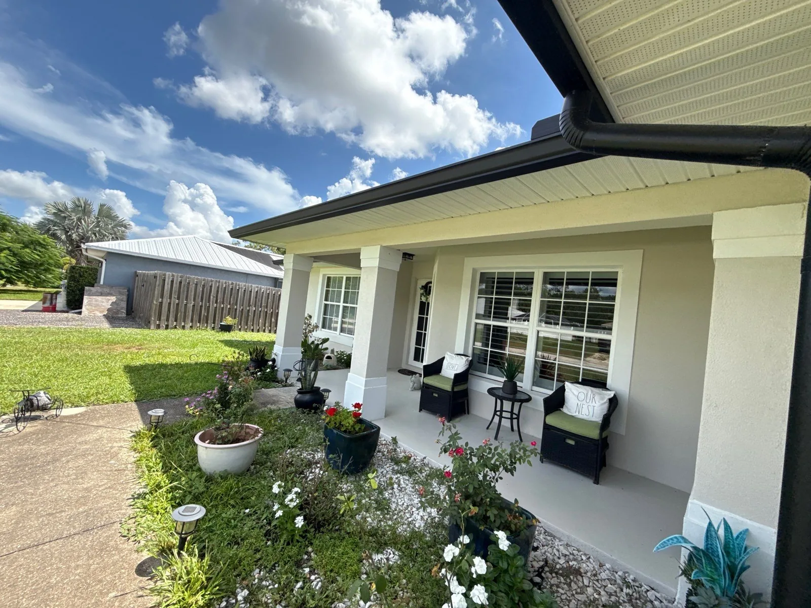 Exterior Painting in Vero Lake Estates, Florida. Front porch of a single-storey home with pale yellow walls, white columns, trim and soffits, patio chairs and flowering landscaping under a blue sky.