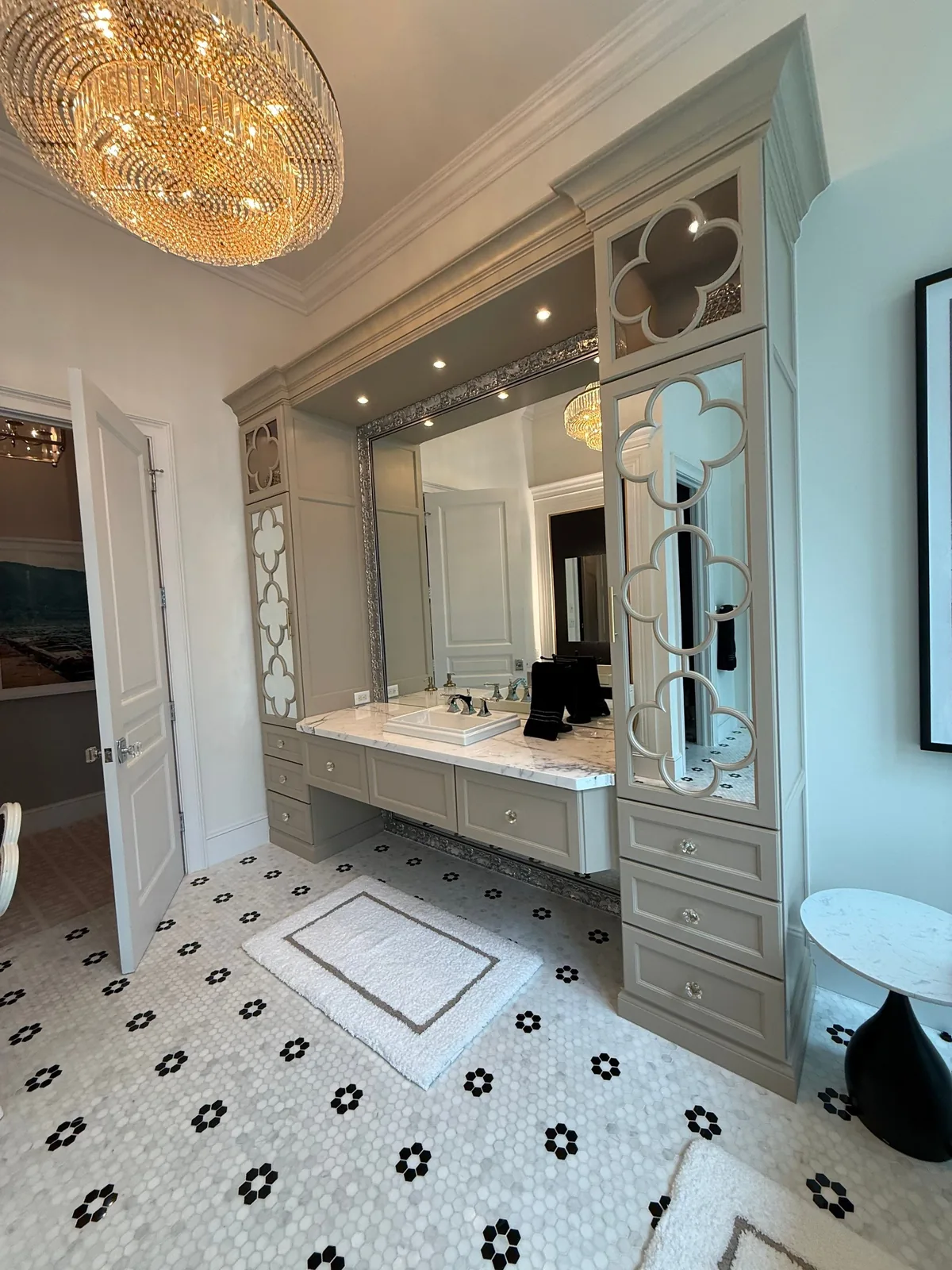Cabinet Refinishing in Wellington, Florida. Bathroom vanity with grey-green painted cabinetry and mirrored fretwork side towers, marble countertop, large mirror, beaded chandelier and black-and-white patterned tile floor.