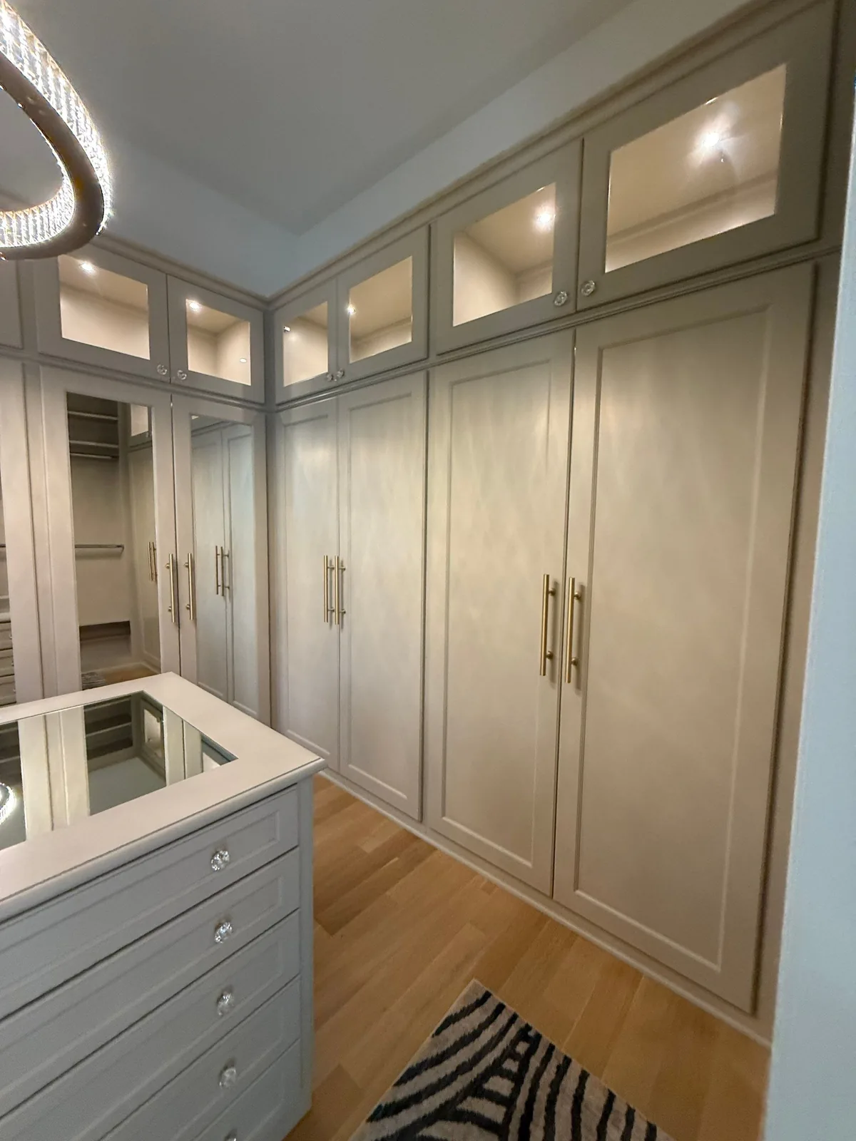 Cabinet Refinishing in Wellington, Florida. Walk-in closet with floor-to-ceiling built-in cabinetry in a light neutral finish, glass-fronted upper doors and brass hardware.
