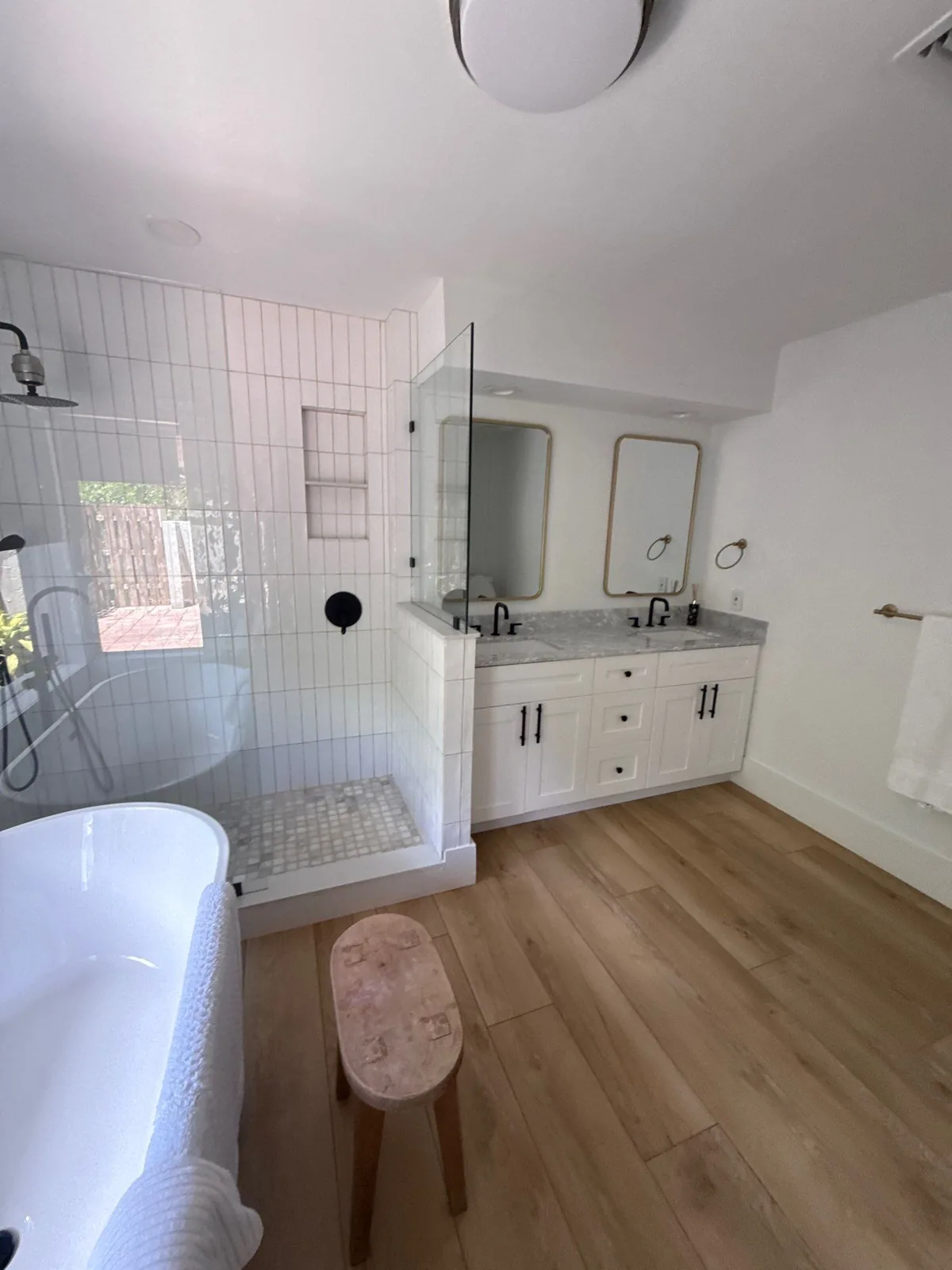 Interior Painting in Wellington, Florida. Finished bathroom with white walls and ceiling, white tiled walk-in shower with glass panel, double vanity with two mirrors and wood-look plank flooring.
