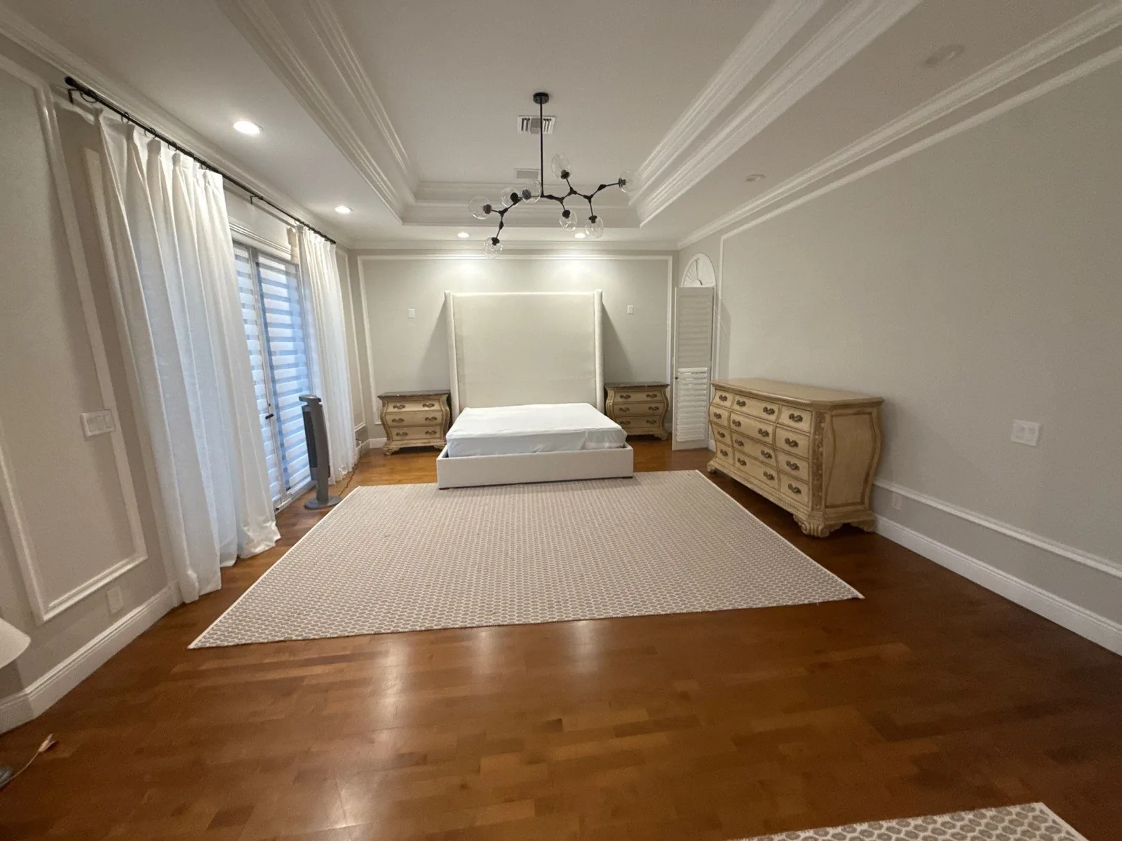 Interior Painting in Wellington, Florida. Large bedroom with a white tray ceiling and crown moulding, pale walls, dark wood floor, bed with upholstered headboard and a wood dresser.
