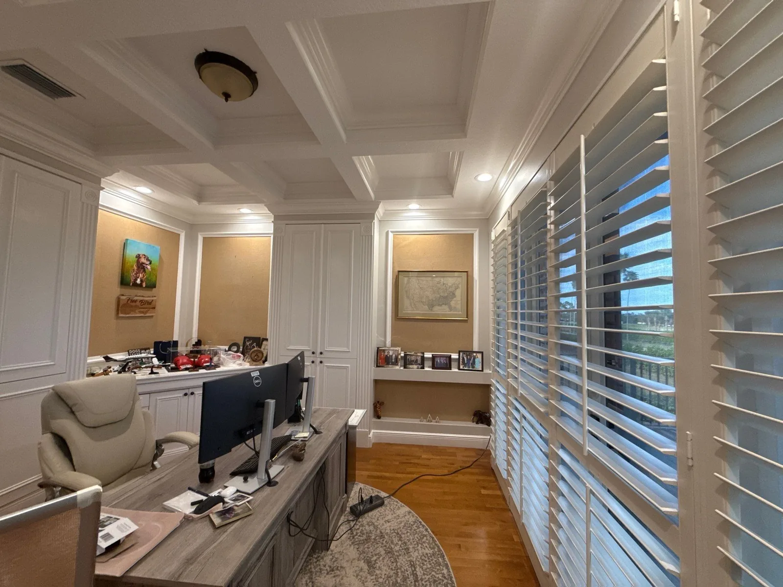 Interior Painting in Wellington, Florida. Home office with a white coffered ceiling, built-in cabinetry and wall panelling, plantation shutters and wood flooring.