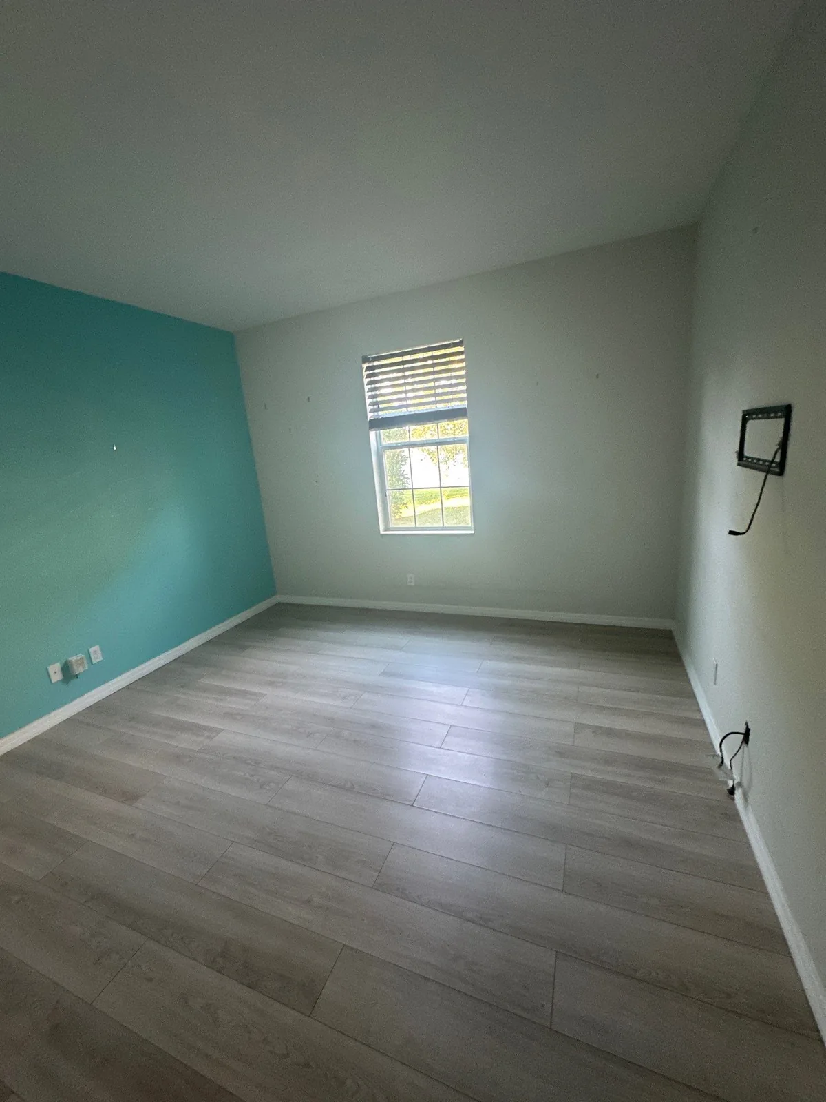Interior Painting in Wellington, Florida. Empty bedroom with a teal accent wall, off-white walls, a blinded window and grey plank flooring.