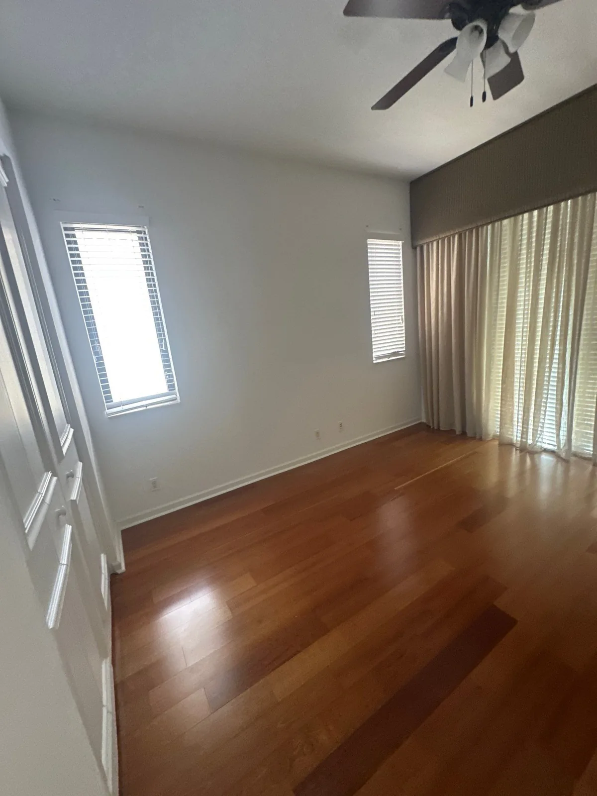 Interior Painting in Whisper Walk, Florida. Empty bedroom with painted light walls and a darker accent wall, white baseboards, two blinded windows, floor-length curtains and hardwood floor.