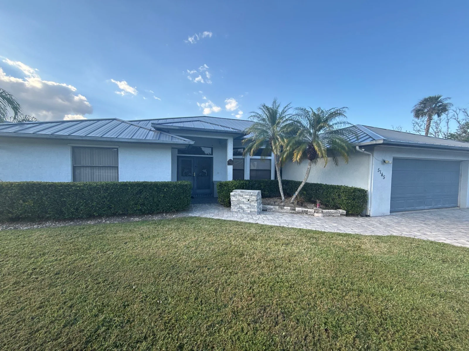 Exterior Painting in White City, Florida. Single-storey house front elevation in light grey with white trim and a blue front door, metal roof, palm trees and lawn.