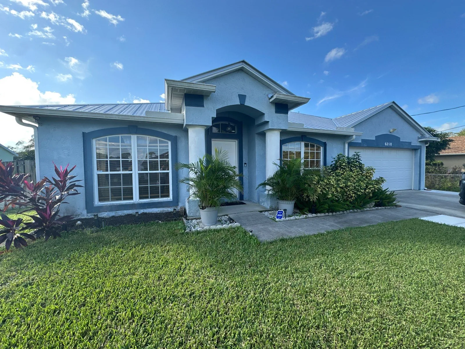 Exterior Painting in White City, Florida. Full front elevation of a single-storey house, blue-grey stucco body with white trim and columns, metal roof, attached garage and green lawn.