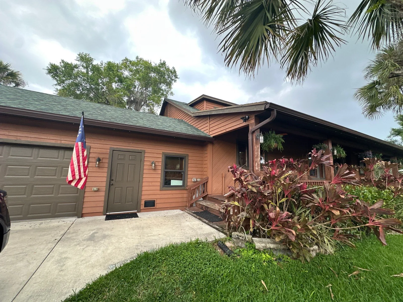 Exterior Painting in White City, Florida. Single-storey home with brown wood siding, brown garage door and shingle roof, with palms and shrubs in front.