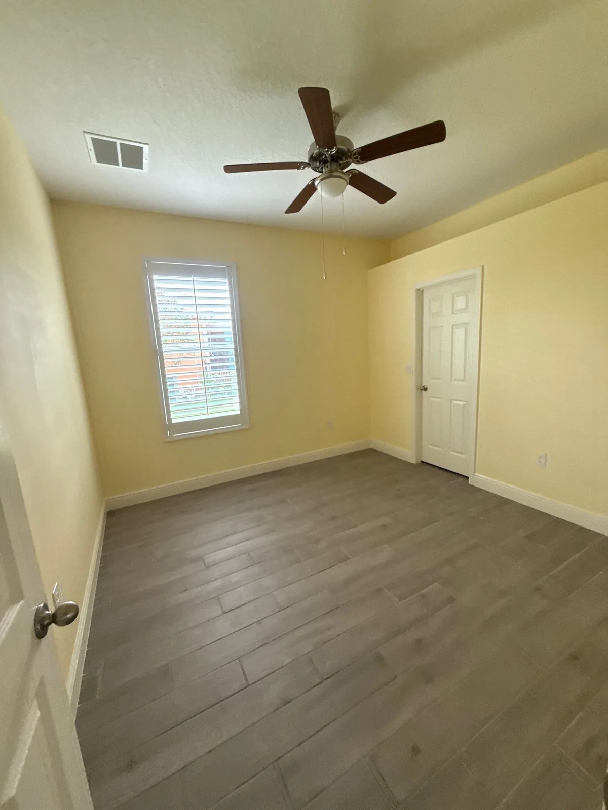 Interior Painting in Winter Beach, Florida. Empty bedroom with freshly painted pale yellow walls, white trim and door, ceiling fan, shuttered window and wood-look plank flooring.